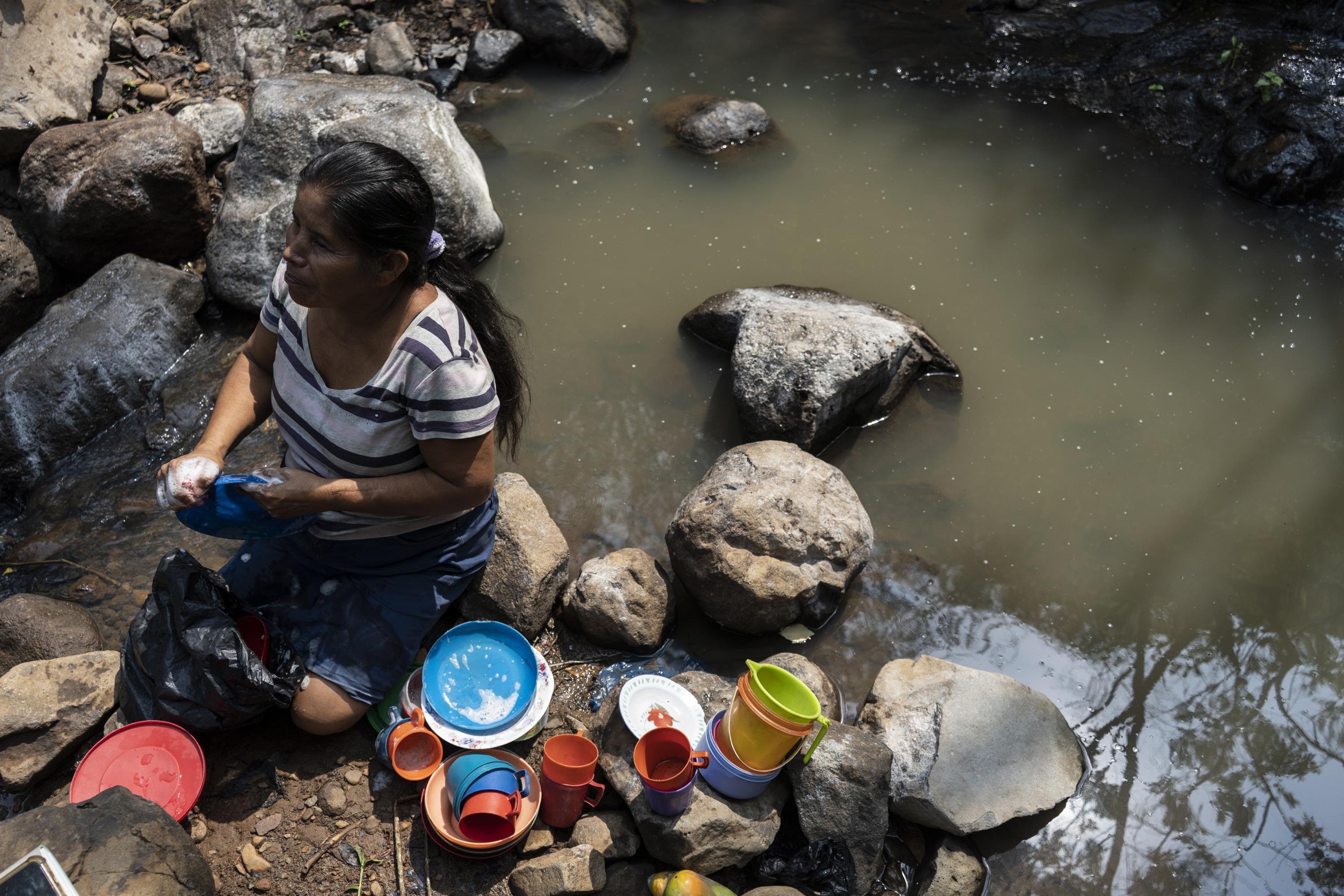 Santos Ángeles Hernández, 63, makes regular trips to El Llano creek to wash her family’s clothes and dishes. They walk through the mountains and along dirt paths for about 15 minutes to reach the murky stream that flows down from the hills and springs up between two large boulders, after passing through most of the pastures that surround the village of Matazano, in the canton of Valle Grande, north of San Simón — one of the poorest and most remote municipalities in the department of Morazán. Santos scrubs and rinses her dishes in the water, puts them in a black plastic bag, and carries them back home. She, her husband, and their four grandchildren eat from these dishes. Santos also makes a living by washing her neighbors’ clothes; she earns about $5 a day, which she spends on the two pounds of beans that will feed her family for two weeks.
